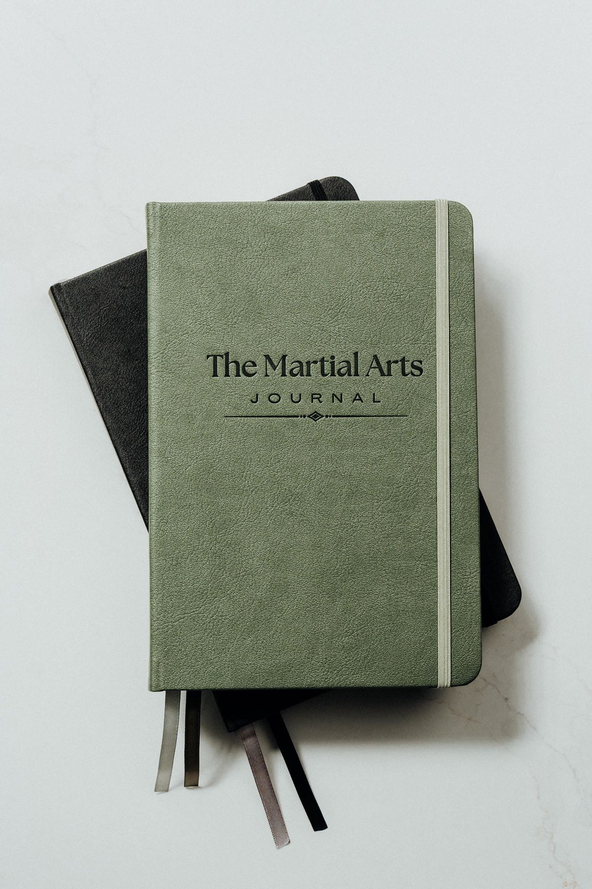 A green hardcover journal titled 'The Martial Arts Journal' stacked on top of a black notebook, with ribbon bookmarks visible, on a light gray background.
