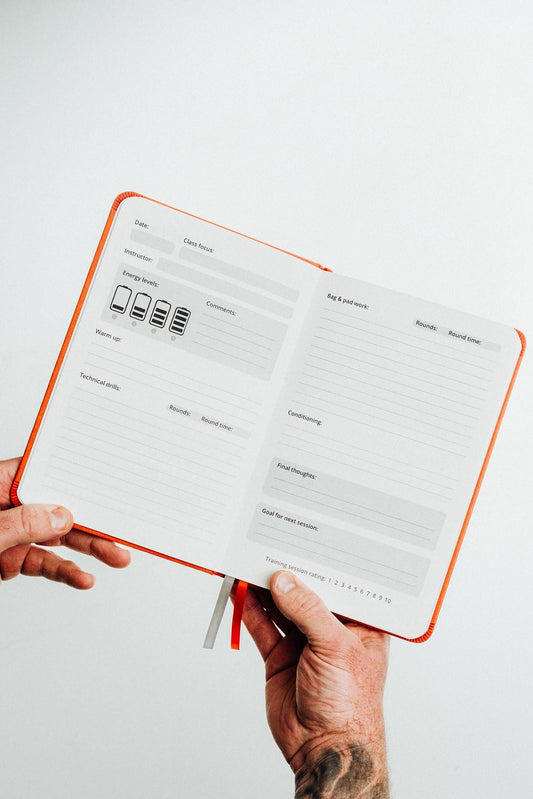 A person's hands holding an open orange-bound planner or notebook with white lined pages against a plain light gray background.