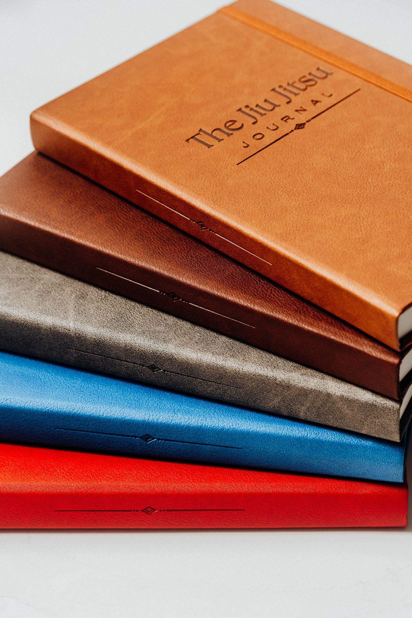 Five journals stacked - light brown on top, dark brown, then grey, blue and red. "The Jiu Jitsu Journal" is written on the top journal