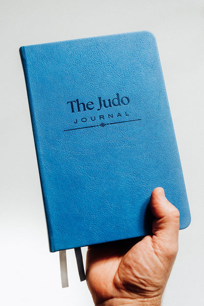 Blue notebook titled 'The Judo Journal' held by a hand against a light background