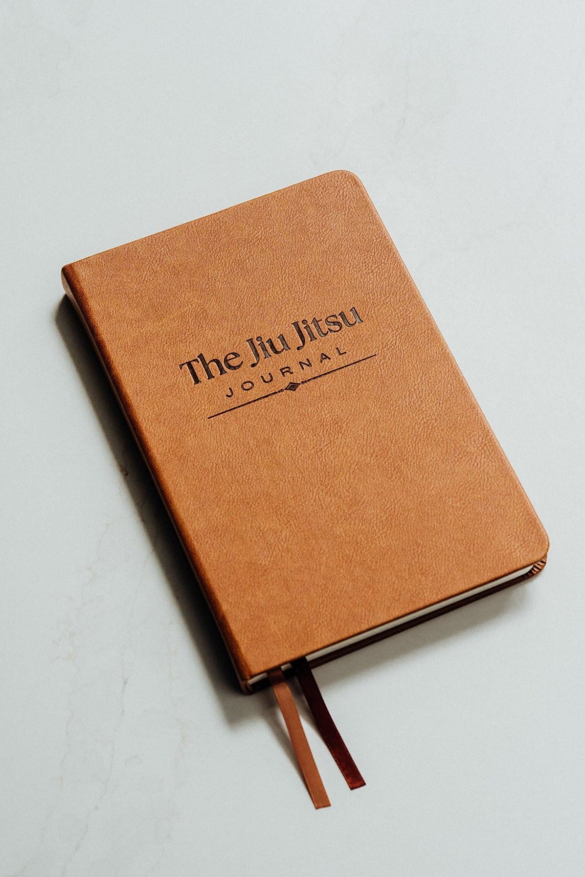 A tan leather-bound journal titled 'The Jiu Jitsu Journal' with a debossed logo, resting on a light gray marble surface with two brown ribbon bookmarks visible.