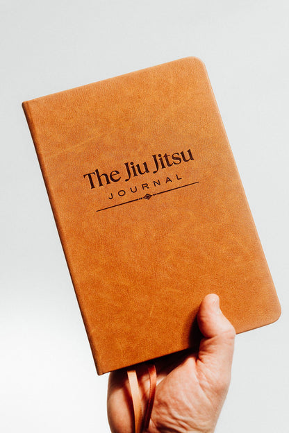 A hand holding a tan leather-bound journal with the title 'The Jiu Jitsu Journal' debossed logo