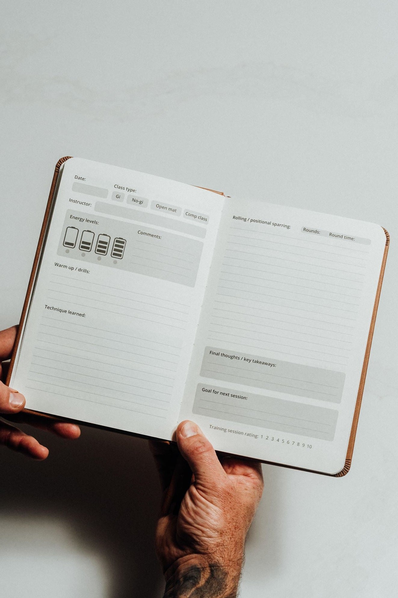 A person's hands holding open a brown leather-bound planner or notebook showing lined daily planning pages with gray boxes and small icons on a light gray background.