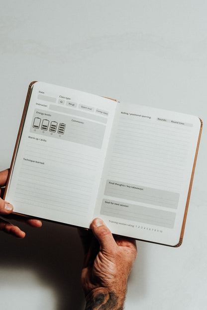 A person's hands holding open a brown leather-bound planner or notebook showing lined daily planning pages with gray boxes and small icons on a light gray background.