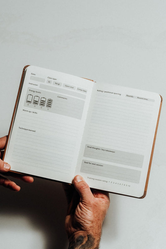 A person's hands holding open a brown leather-bound planner or notebook showing lined daily planning pages with gray boxes and small icons on a light gray background.