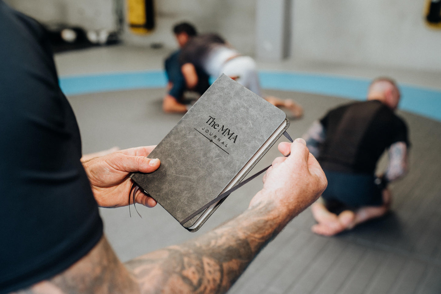 Person holding a grey notebook with 'The MMA Journal' logo in a gym setting.
