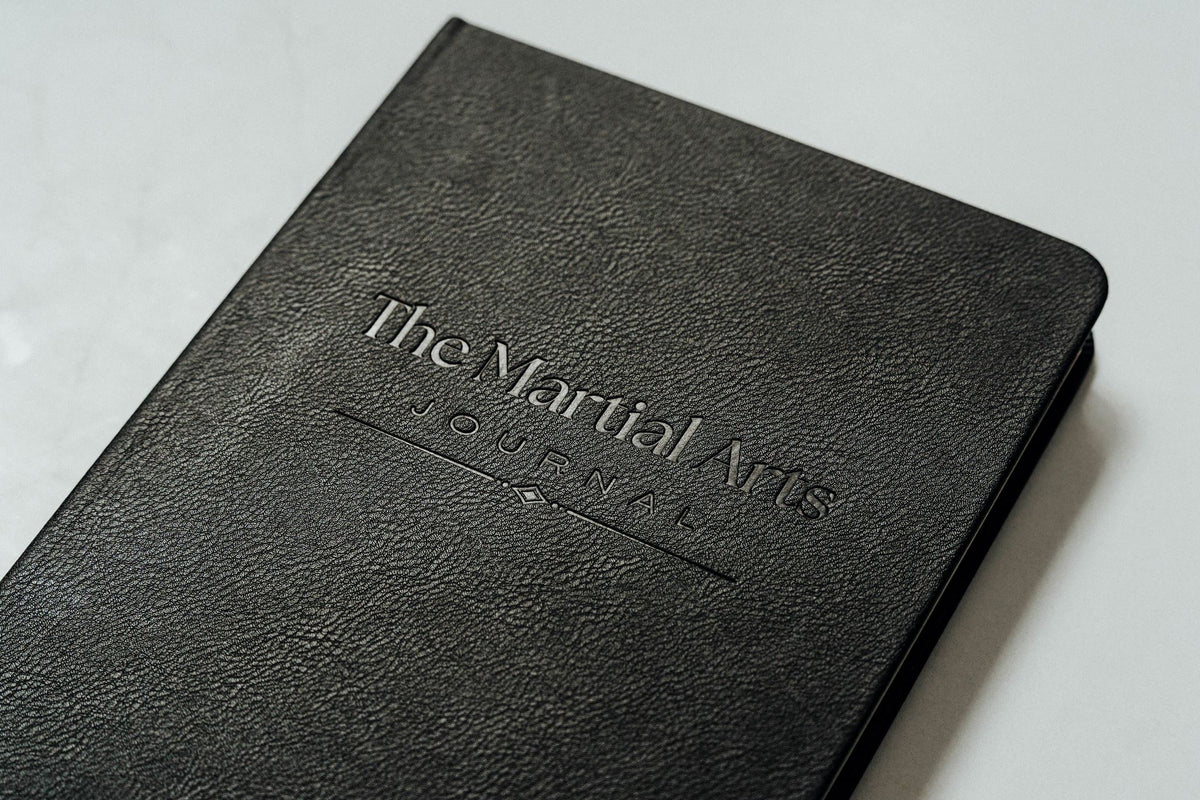 A close-up of a black textured leather journal with the title 'The Martial Arts Journal' embossed on the cover, resting on a light gray surface.