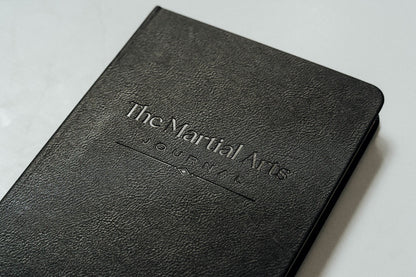 A close-up of a black textured leather journal with the title 'The Martial Arts Journal' embossed on the cover, resting on a light gray surface.