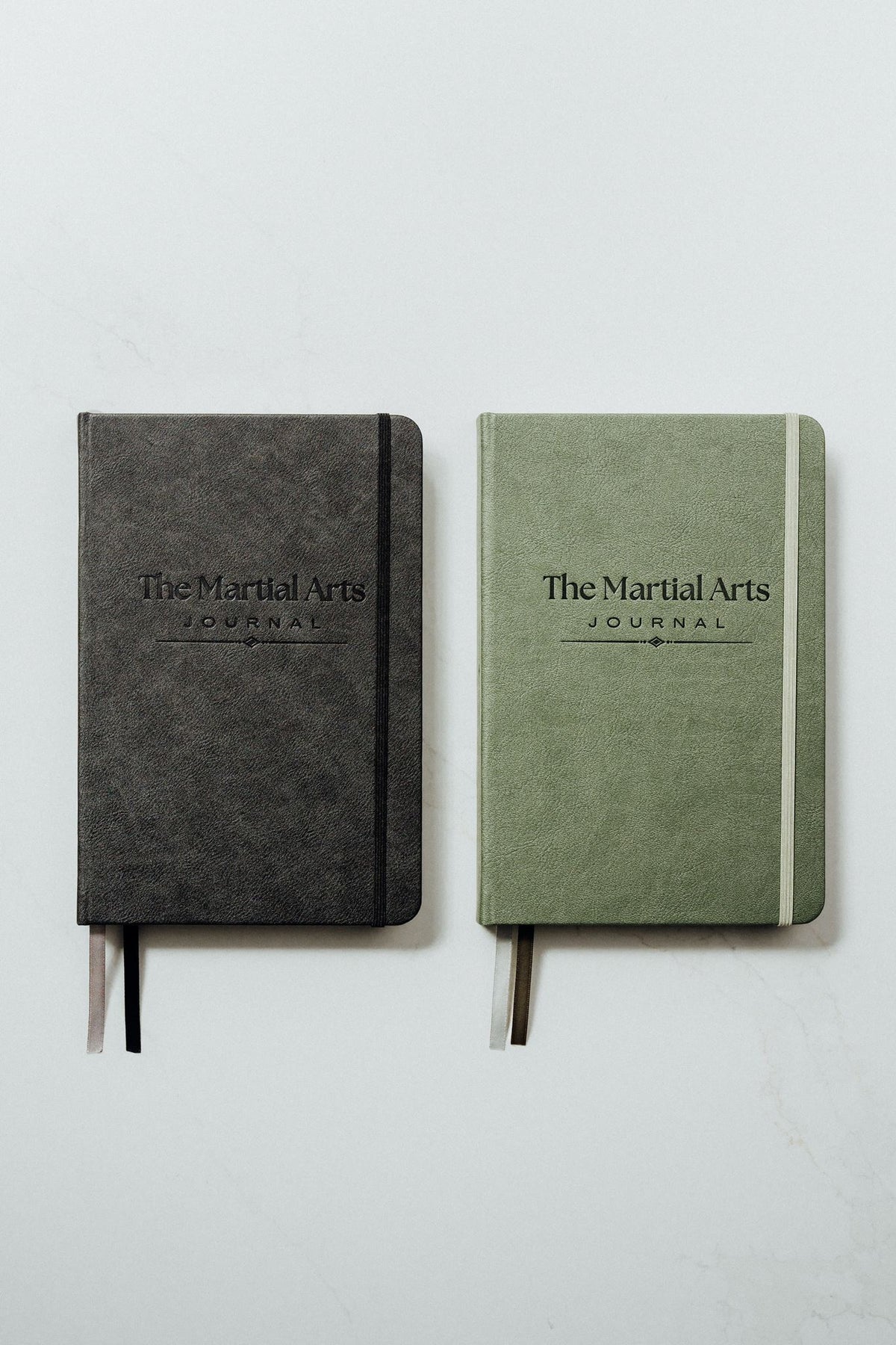 The martial arts journals side by side on a light gray background. The left journal is black with a black elastic closure, and the right journal is sage green with a green elastic closure.