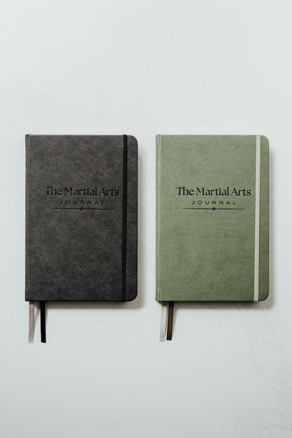 The martial arts journals side by side on a light gray background. The left journal is black with a black elastic closure, and the right journal is sage green with a green elastic closure.