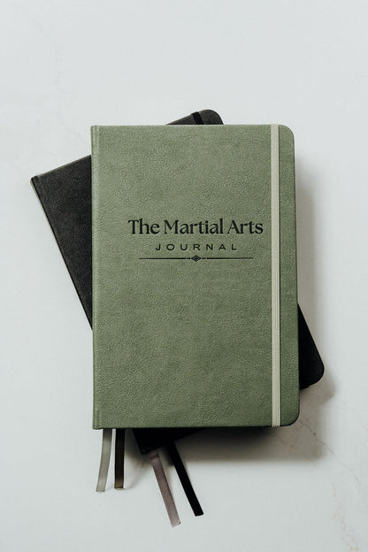 A green hardcover journal titled 'The Martial Arts Journal' stacked on top of a black notebook, with ribbon bookmarks visible, on a light gray background.