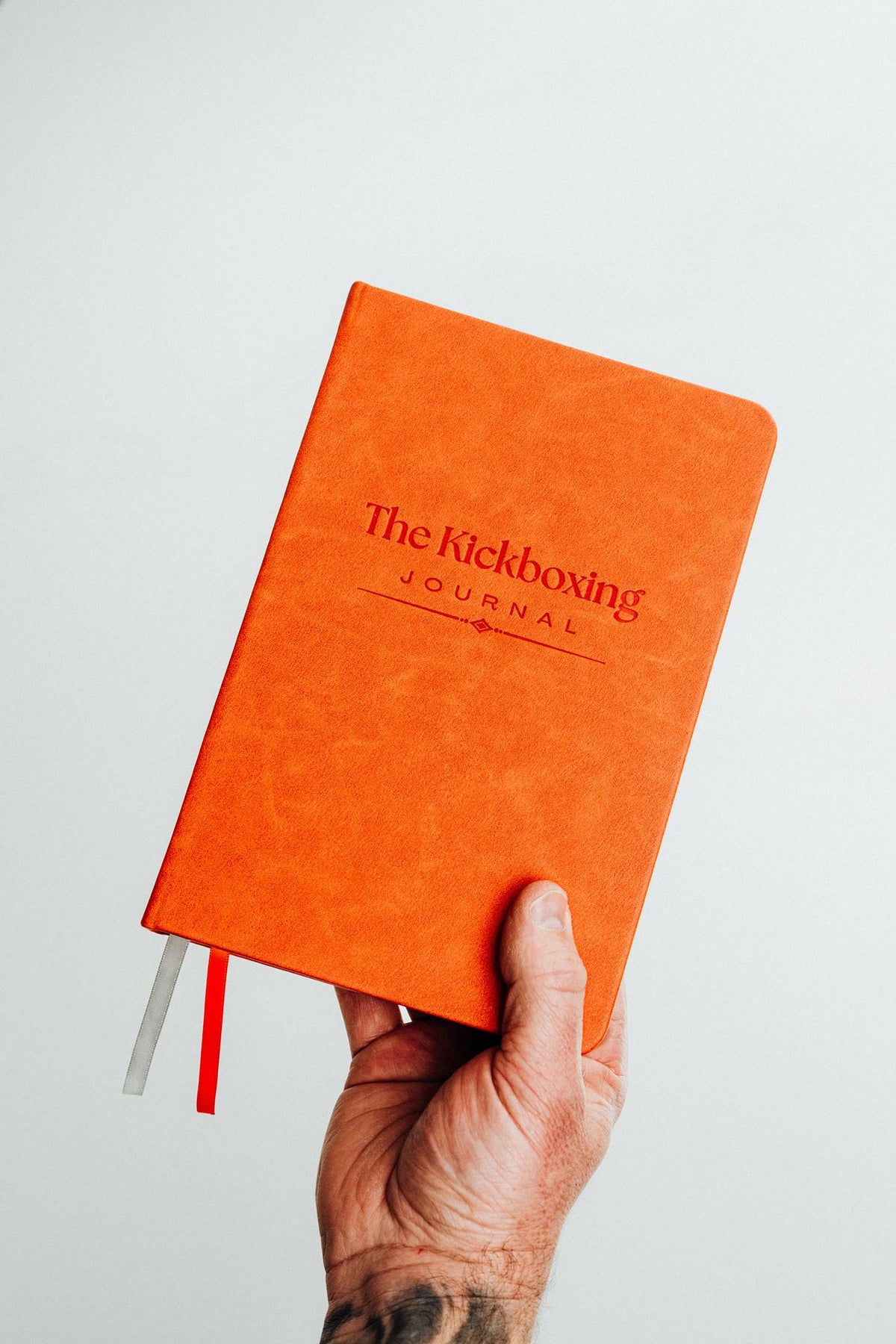 A hand holding an orange hardcover journal titled 'The Kickboxing Journal' against a plain light gray background.
