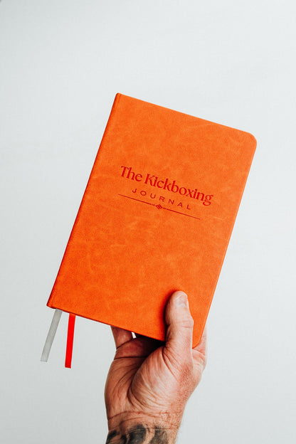 A hand holding an orange hardcover journal titled 'The Kickboxing Journal' against a plain light gray background.