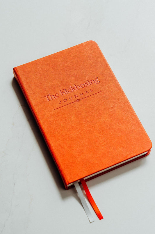 Orange notebook with 'The Kickboxing Journal' debossed on the cover on a light grey background