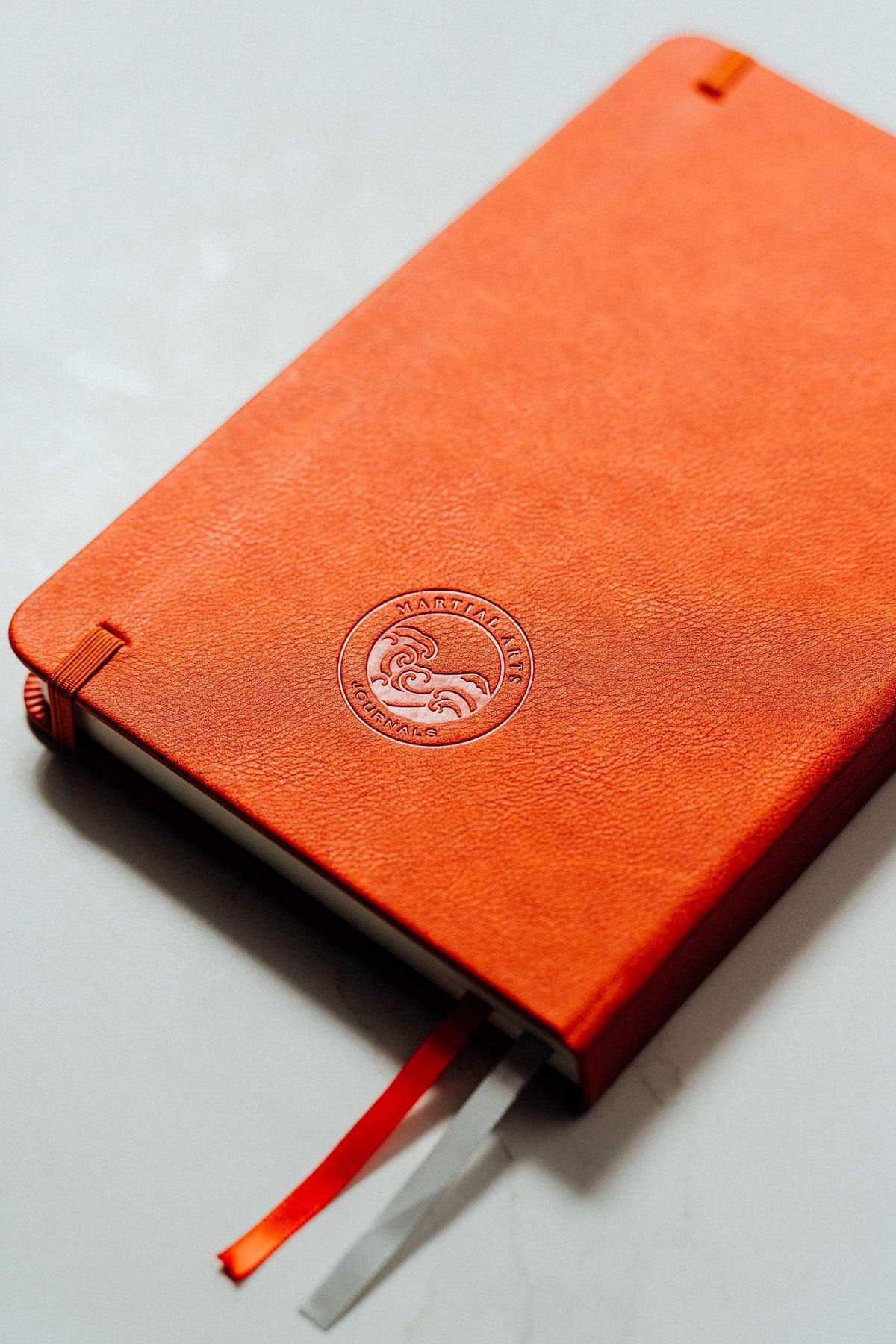 An orange textured leather notebook with a debossed circular logo on the back cover, resting on a light gray surface with an orange and silver ribbon bookmark.