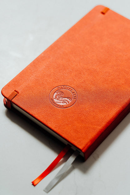 An orange textured leather notebook with a debossed circular logo on the back cover, resting on a light gray surface with an orange and silver ribbon bookmark.