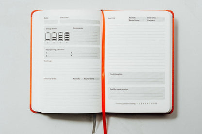An open planner or notebook with ivory pages, orange binding, and a matching orange ribbon bookmark, photographed from above on a light gray background. This is the main kickboxing sparring pages