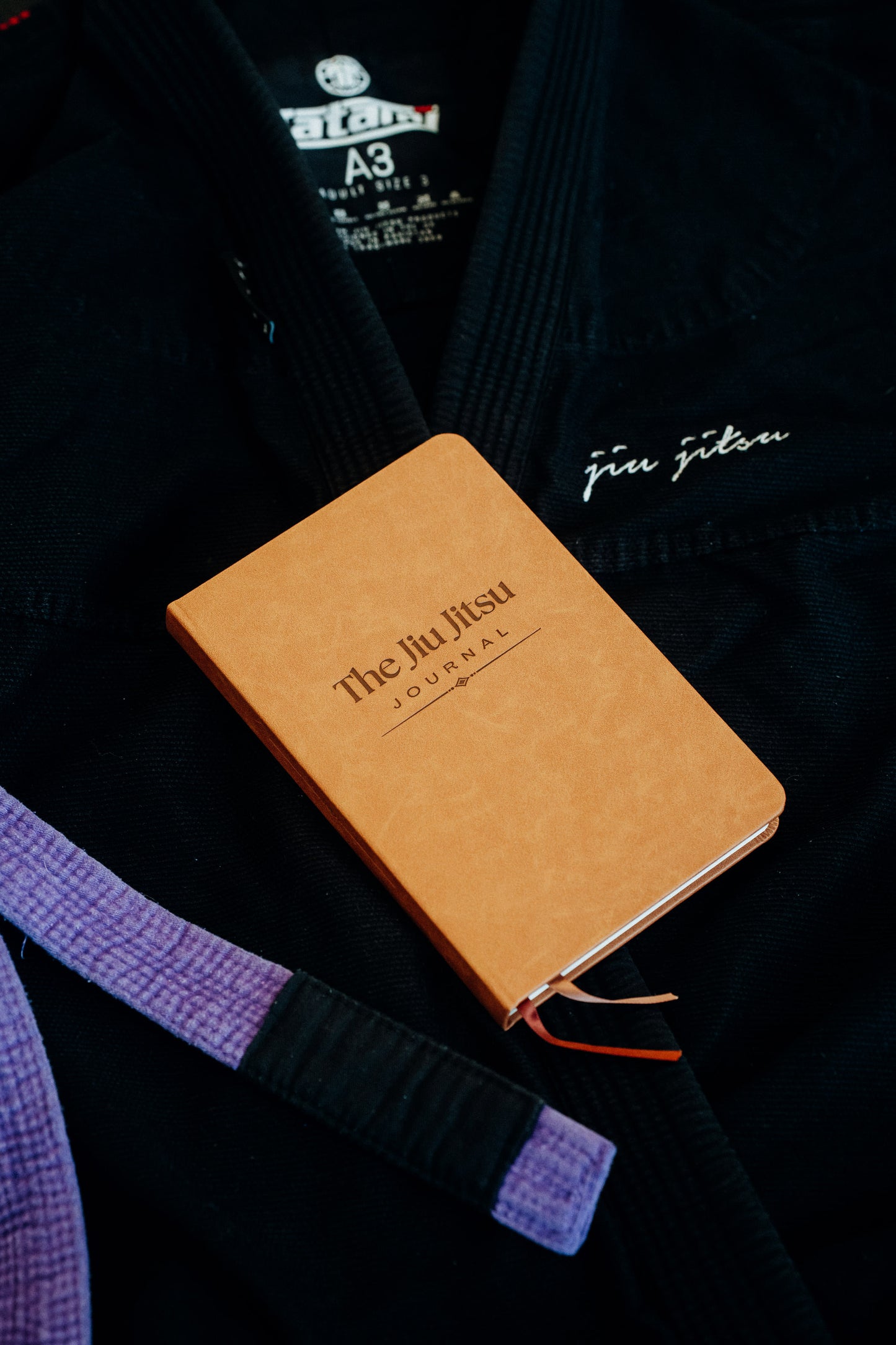 Brown notebook with 'The Jiu Jitsu Journal' on a black gi with a purple belt.